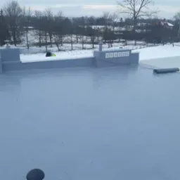 Płaski dach pokryty szarą, jednolitą powłoką hydroizolacyjną, z widocznymi elementami wentylacyjnymi i zwiniętą rolką papy, w tle zimowy krajobraz z drzewami i domami.