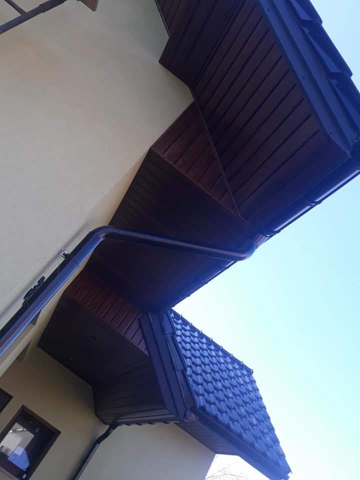 Fragment dachu z ciemnej dachówki ceramicznej i drewnianą podbitką w Zduńskiej Woli, widoczne orynnowanie i elewacja budynku w kremowym odcieniu.