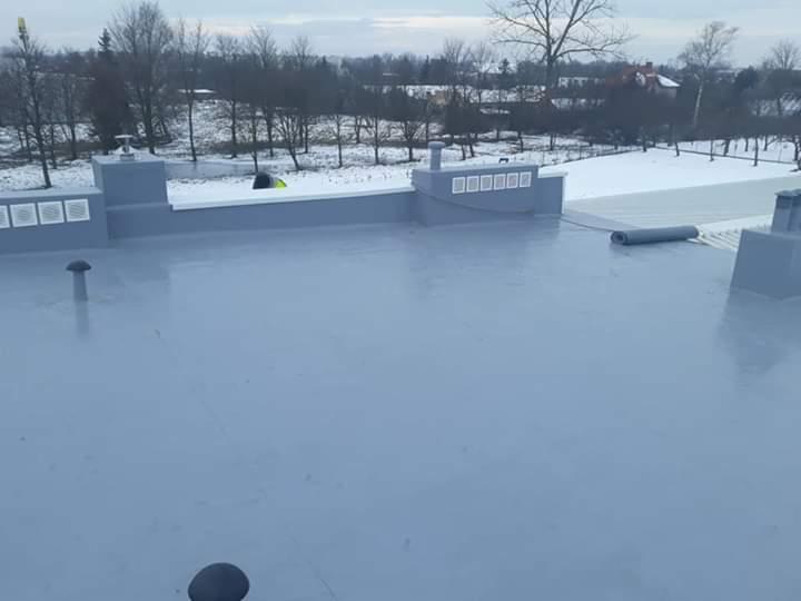 Płaski dach pokryty szarą, jednolitą powłoką hydroizolacyjną, z widocznymi elementami wentylacyjnymi i zwiniętą rolką papy, w tle zimowy krajobraz z drzewami i domami.