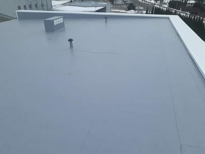 Płaski, szary dach pokryty papą termozgrzewalną z widocznymi kominkami wentylacyjnymi i obróbkami blacharskimi, widok z góry w pochmurny dzień.