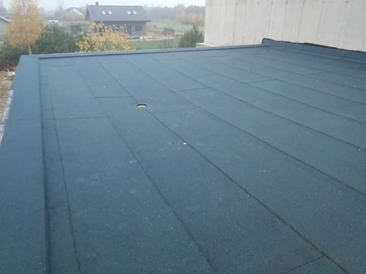 Płaski dach pokryty ciemną papą bitumiczną z widocznym otworem wentylacyjnym i łączeniami, w tle dom i drzewa w pochmurny dzień.