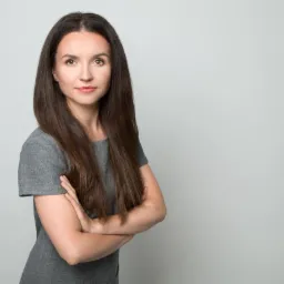 Portret kobiety z długimi, ciemnymi włosami, ubranej w szarą sukienkę, z założonymi rękami na neutralnym, jasnoszarym tle.