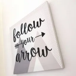 Płótno z nadrukiem 'follow your arrow' w czcionce ozdobnej, z motywem strzał i gór w odcieniach szarości, zawieszone na ścianie.