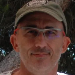 Portret mężczyzny w okularach i czapce z naszywką, uśmiechającego się do kamery na tle drzewa.