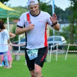 Mężczyzna w sportowym stroju z numerem 138 biegnie po trawie, machając ręką w geście powitania.