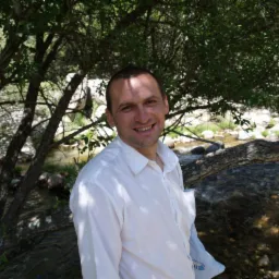 Uśmiechnięty mężczyzna w białej koszuli, na tle strumienia i drzew, w naturalnym otoczeniu. Zdjęcie wykonane w plenerze, z wykorzystaniem naturalnego światła.