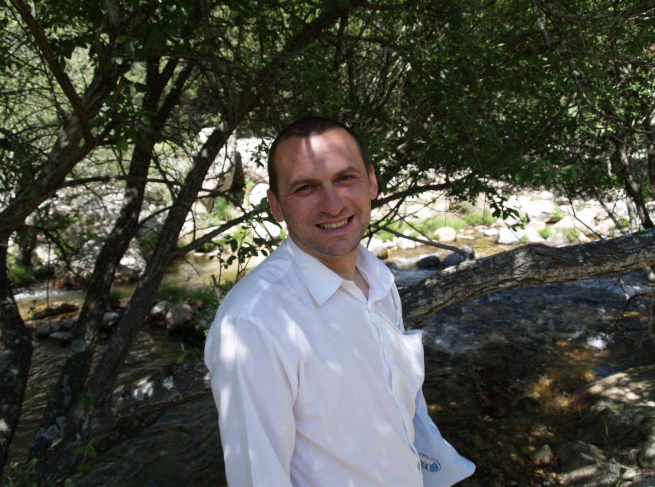 Uśmiechnięty mężczyzna w białej koszuli, na tle strumienia i drzew, w naturalnym otoczeniu. Zdjęcie wykonane w plenerze, z wykorzystaniem naturalnego światła.