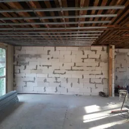 Wnętrze w trakcie prac murarskich: ściana z białych bloczków z widoczną zaprawą, metalowa konstrukcja podwieszanego sufitu, okno z żaluzjami oraz stos profili na podłodze.