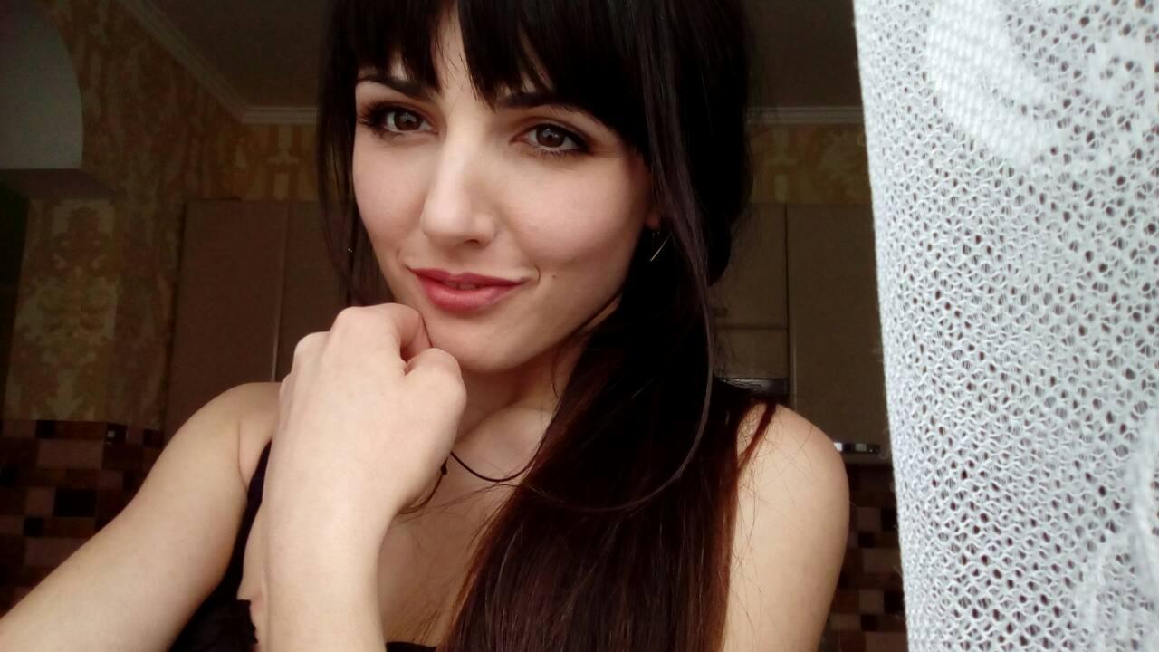 Portret uśmiechniętej kobiety z ciemnymi włosami i grzywką, trzymającej dłoń przy twarzy, z widokiem na wnętrze pomieszczenia przez okno z firanką w tle.