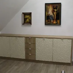 Długa komoda z frezowanymi drzwiami w geometryczne wzory, trzy szuflady pośrodku, drewniany blat, w tle skośna ściana poddasza z dwoma obrazami.