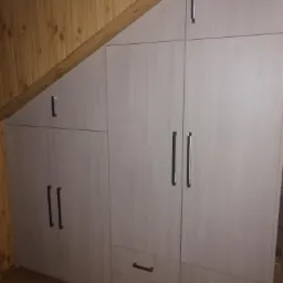 Szara szafa wnękowa na wymiar, z dopasowaniem do skosu dachu, z podwójnymi uchwytami i małymi szufladami na dole, ustawiona na brązowych płytkach.