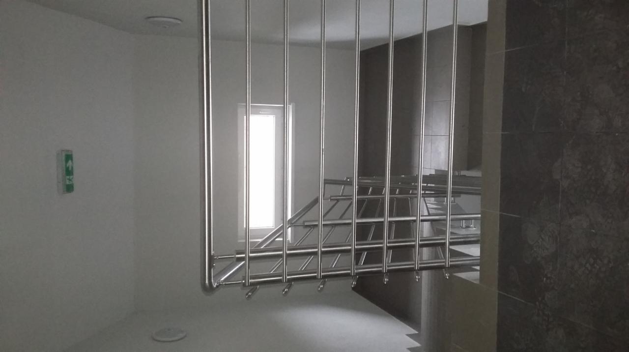 Nowoczesna, metalowa balustrada schodowa z pionowymi prętami, widziana od dołu, z widocznym znakiem ewakuacyjnym na ścianie i oknem w tle.