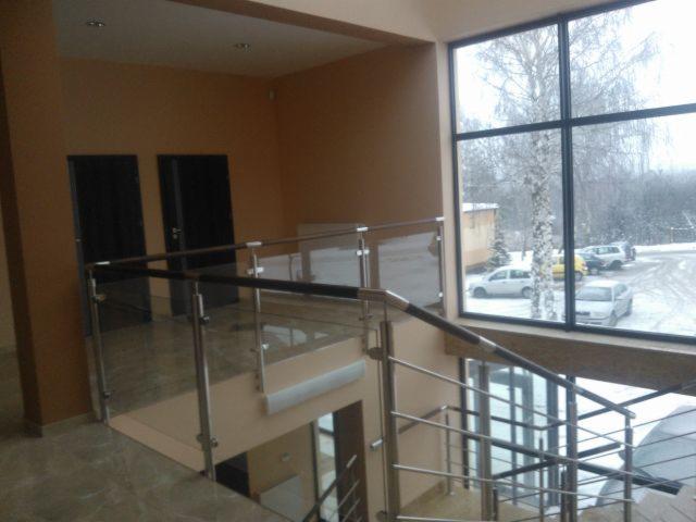 Wnętrze domu w trakcie rozbudowy: widok na klatkę schodową z balustradą ze stali nierdzewnej i szkła, duże okno z widokiem na zimowy krajobraz.