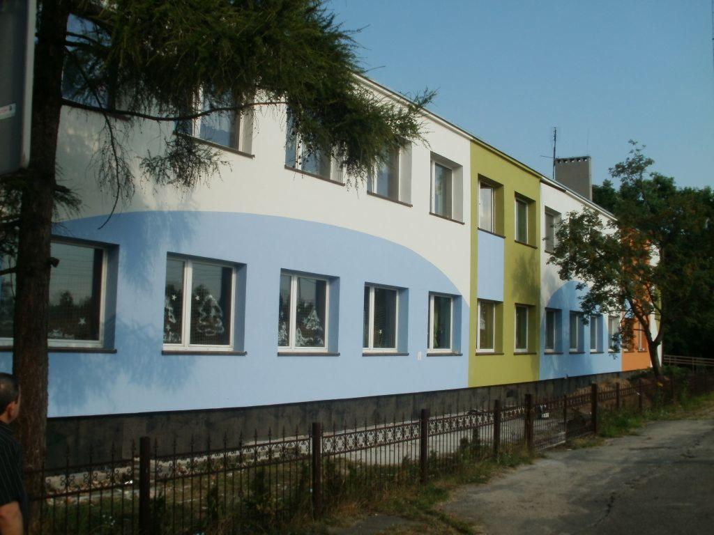 Elewacja budynku po rozbudowie z zastosowaniem kolorowej palety: błękitu, bieli, zieleni i pomarańczu, widoczne okna z dekoracjami świątecznymi.