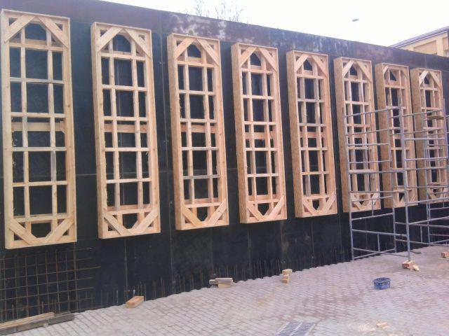 Montaż drewnianych ram okiennych z ozdobnymi trójkątami na czarnej ścianie zewnętrznej budynku w trakcie rozbudowy, widoczne rusztowania i materiały budowlane.