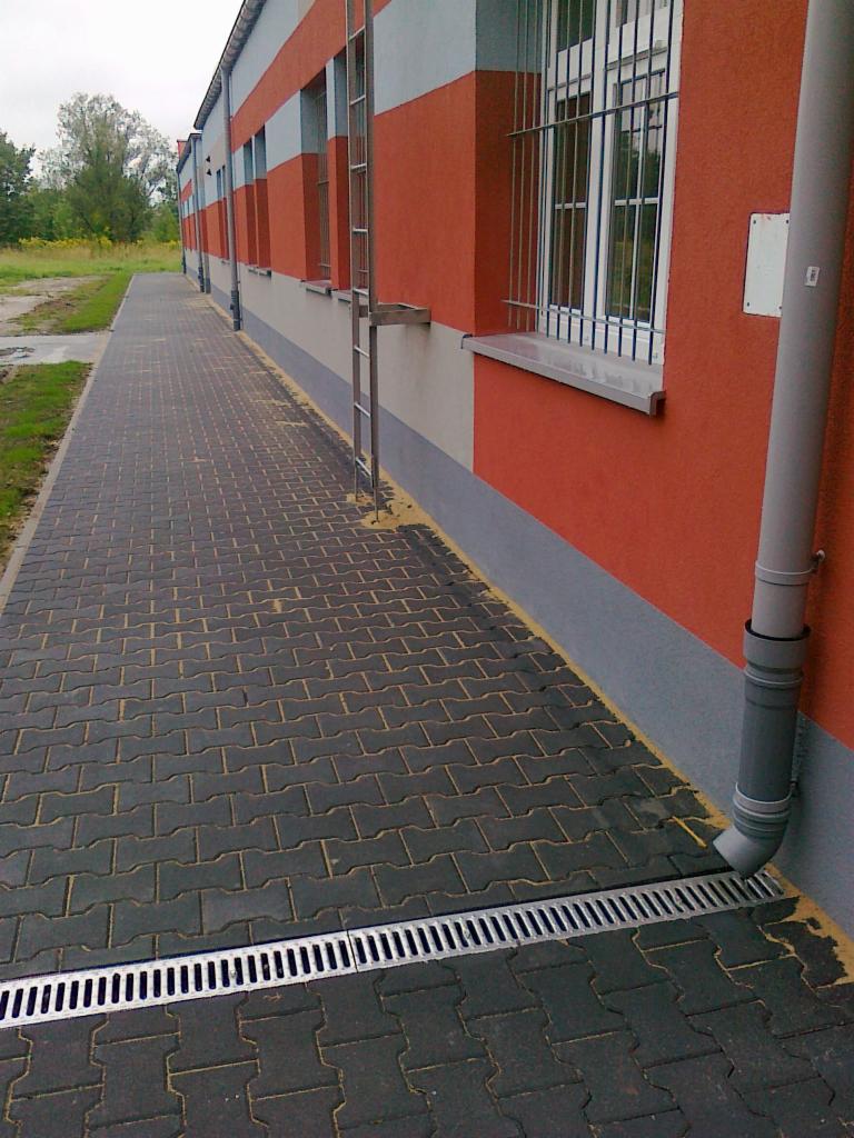 Elewacja budynku z pomarańczowo-szarymi pasami, chodnik z szarej kostki brukowej i kratka odpływowa wzdłuż ściany, widoczna drabina przymocowana do budynku oraz rura spustowa.