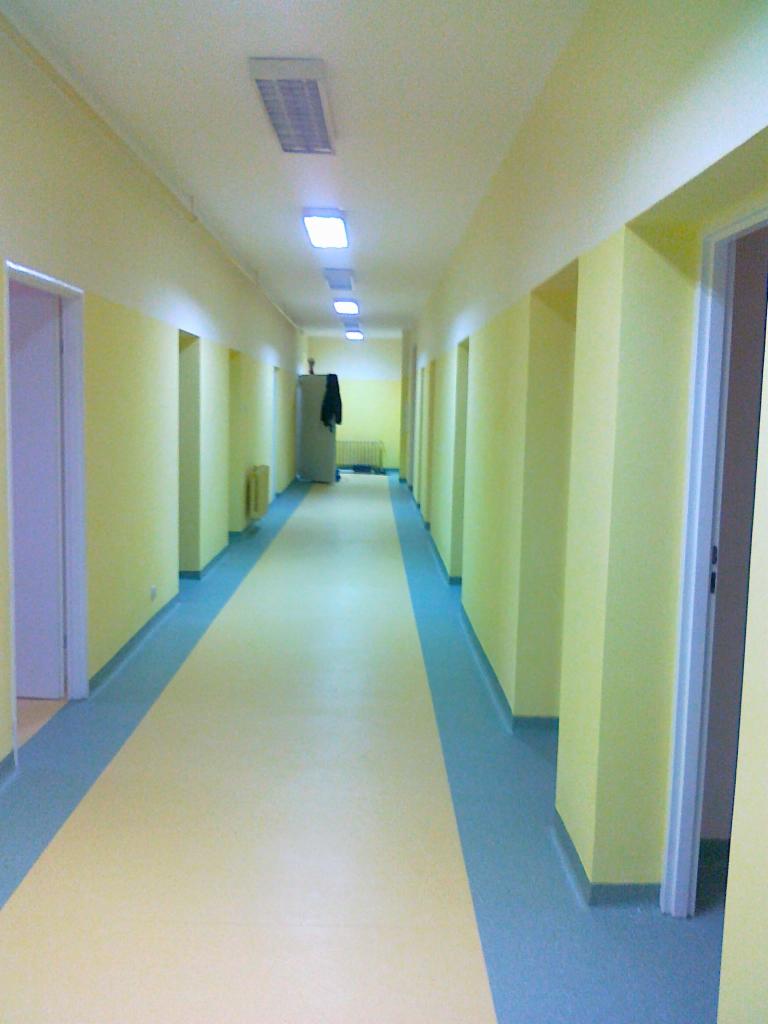 Długi korytarz z żółtymi ścianami i jasną podłogą z niebieskimi pasami, oświetlony rzędami lamp sufitowych, z widocznymi drzwiami po obu stronach i grzejnikiem w oddali.