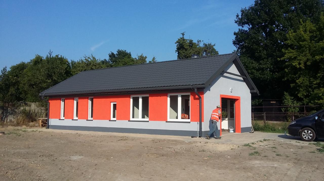 Parterowy budynek z szarą dachówką i pomarańczowo-szarą elewacją; pracownik w kamizelce odblaskowej wchodzi do środka.