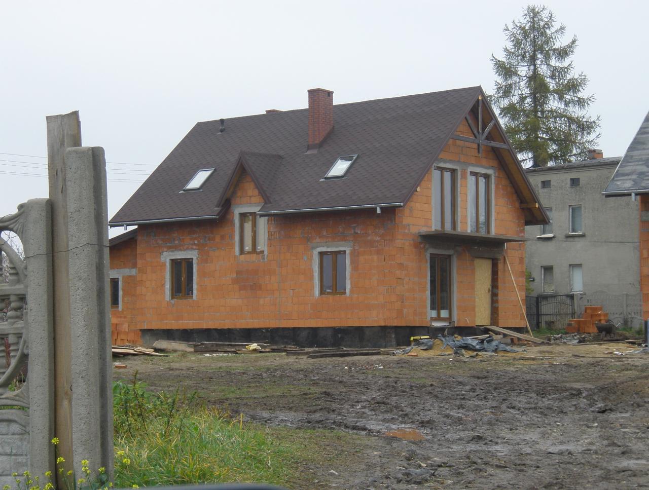 Dom w trakcie budowy z czerwonej cegły, z oknami i szarym dachem, na zewnątrz błotniste podłoże i częściowo wykończony betonowy płot.