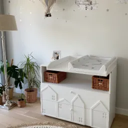 Biała komoda z przewijakiem dla niemowląt, z motywem domków na frontach szuflad, uzupełniona wiklinowymi koszami i dekoracjami ściennymi z motywem balonu i samolotu, w pokoju dziecięcym.