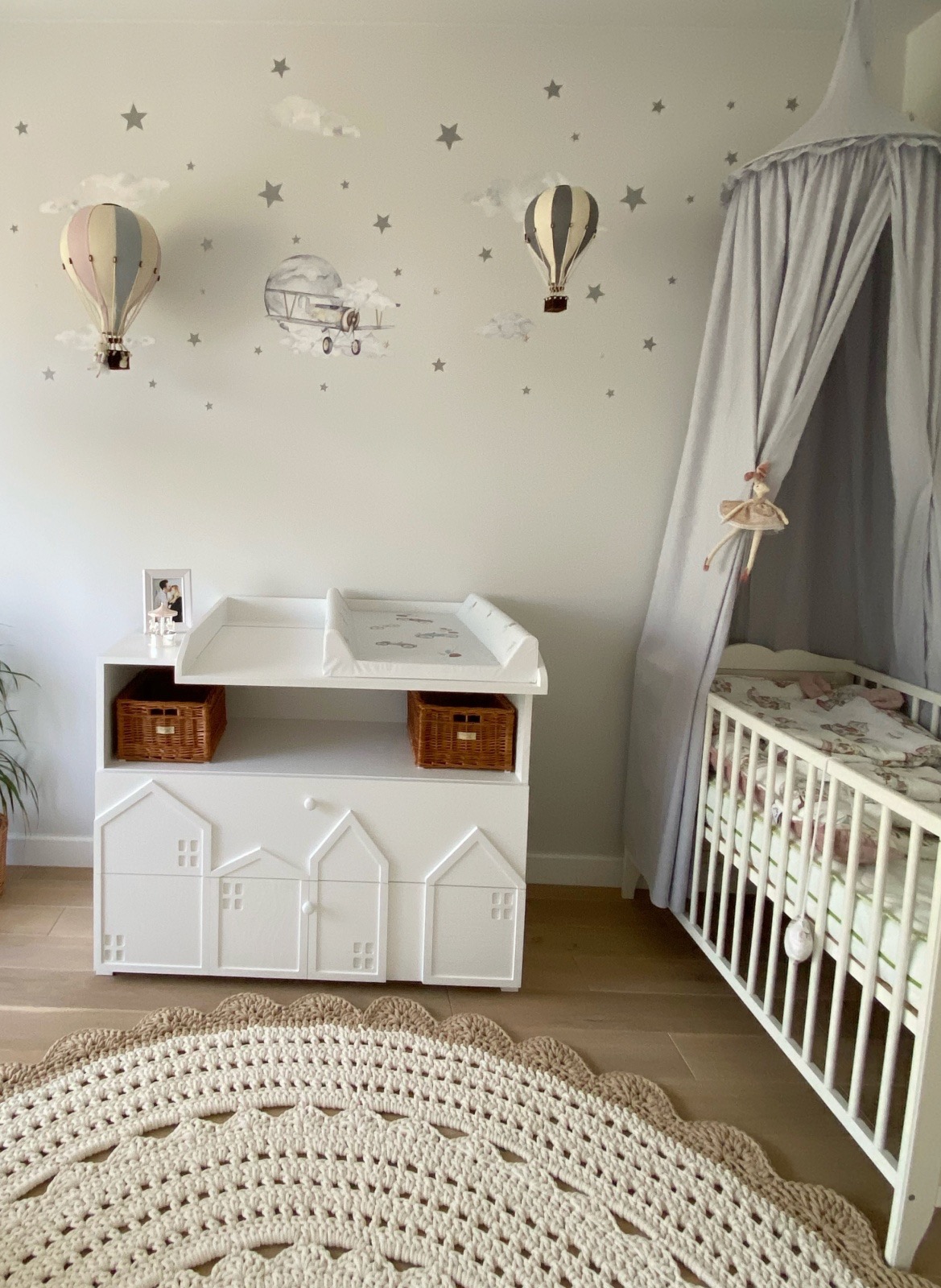 Pokój dziecięcy z białym przewijakiem z motywem domków, łóżeczkiem z baldachimem i dywanem z włóczki, dekoracje ścienne z motywem balonów i samolotu.