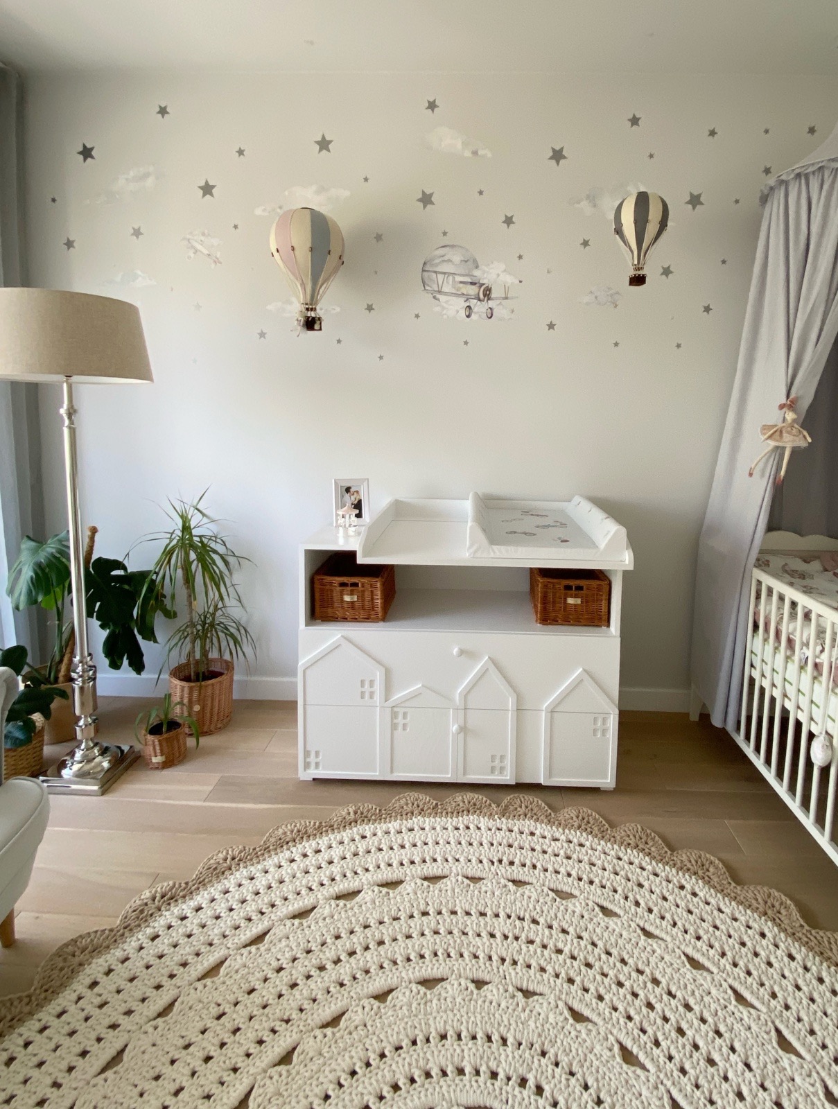Pokój dziecięcy z białym przewijakiem z motywem domków, okrągłym, kremowym dywanem, łóżeczkiem z baldachimem i naklejkami na ścianie z motywem balonów i samolotów.