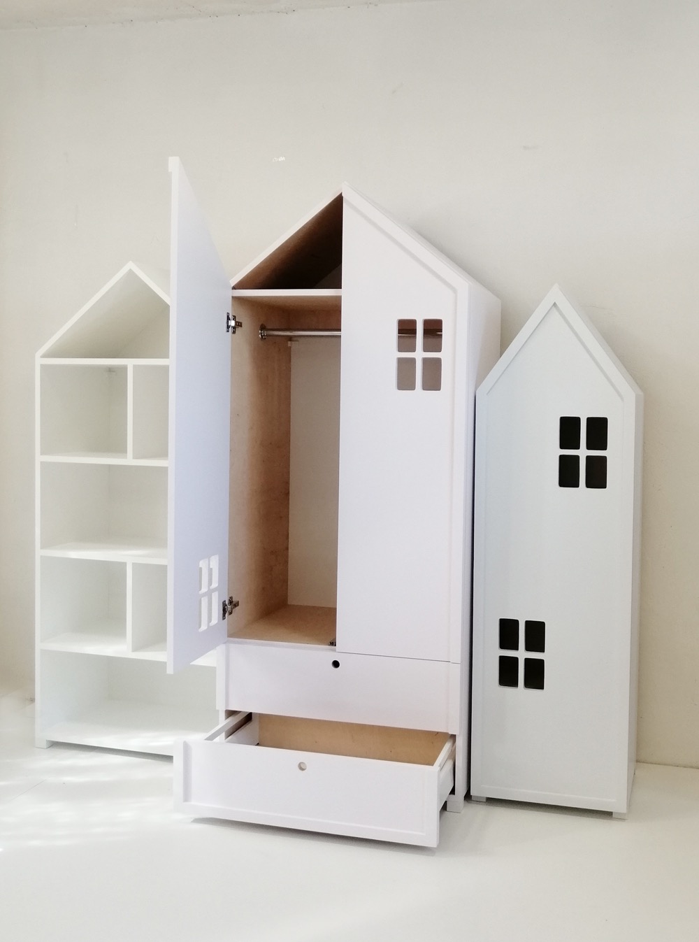 Biała szafa dziecięca z szufladą i półkami, stylizowana na domek z okienkami, z otwartymi drzwiami i wysuniętą szufladą, na jasnym tle.