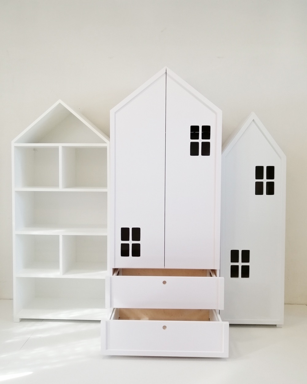 Białe, dziecięce meble w kształcie domków: szafa dwudrzwiowa z wysuwanymi szufladami i regał z półkami, ustawione na białym tle.
