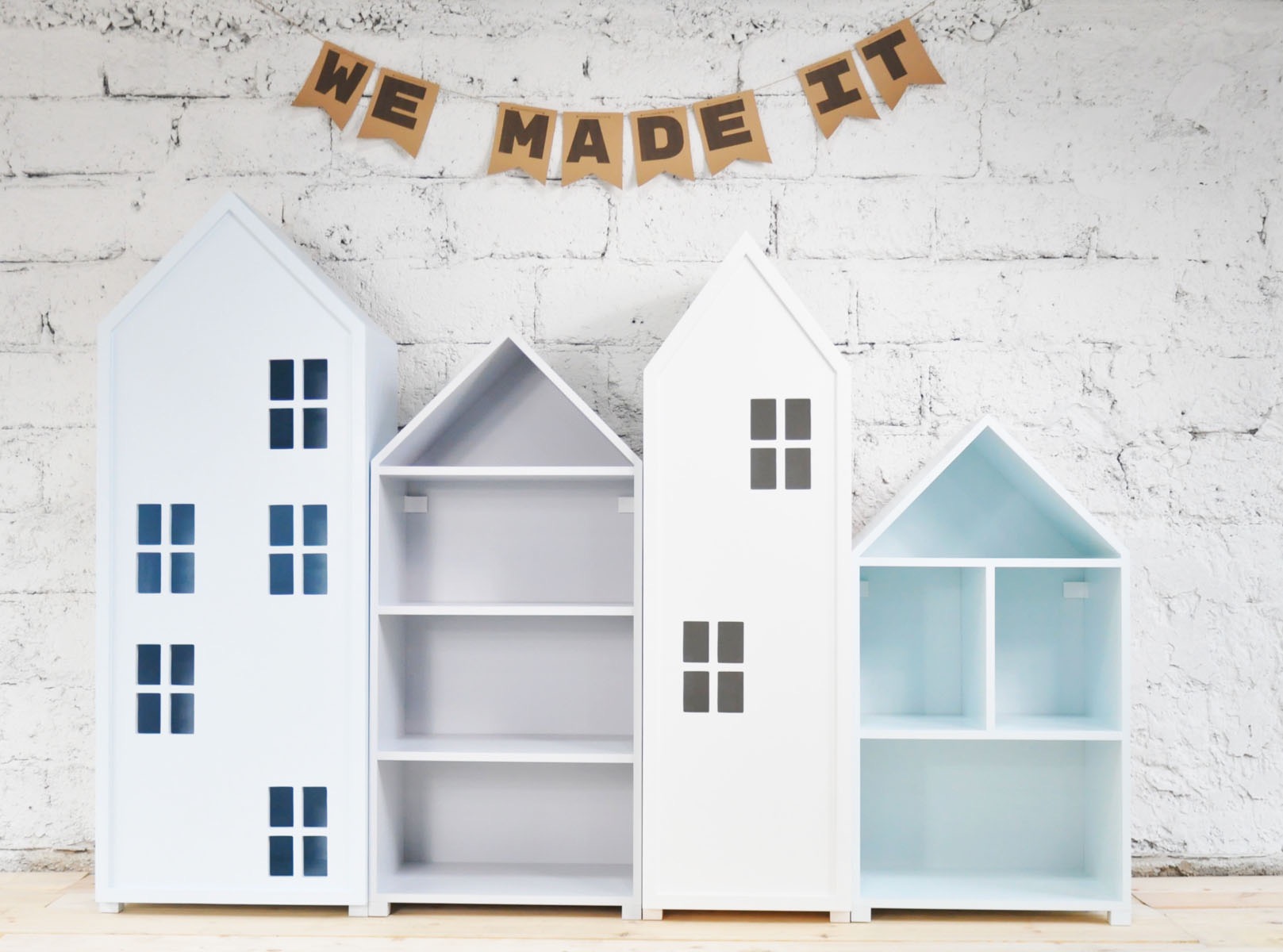 Trzy białe regały dziecięce w kształcie domków, o różnej wysokości i odcieniach błękitu, stoją na drewnianej podłodze na tle białej ceglanej ściany, a nad nimi wisi transparent z napisem 'WE MADE IT'.