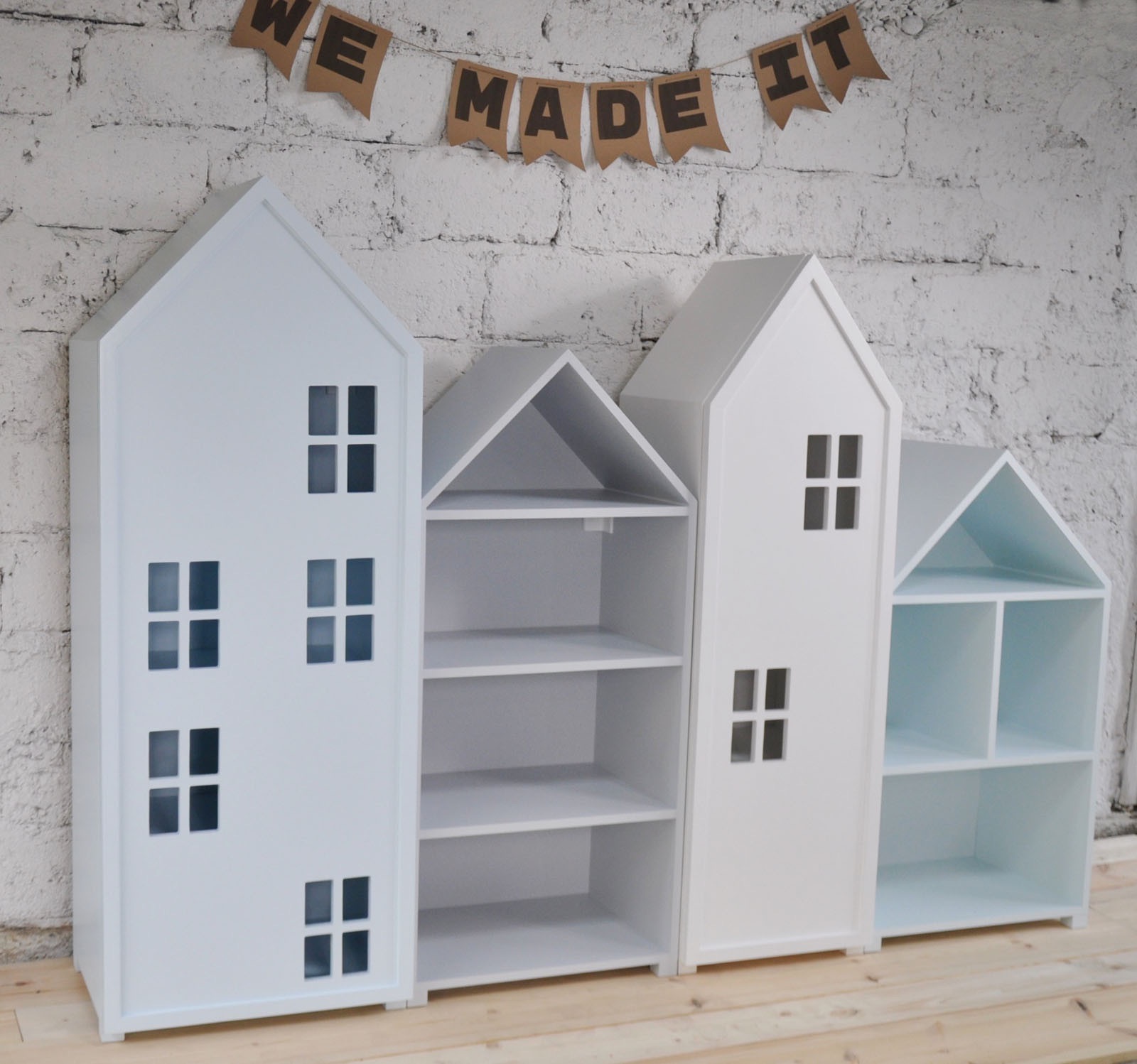 Trzy białe regały w kształcie domków, z 'oknami' i półkami, ustawione obok siebie na drewnianej podłodze w Brzuśniku, z dekoracyjnym napisem 'WE MADE IT' na sznurku wiszącym na tle ściany z białej...