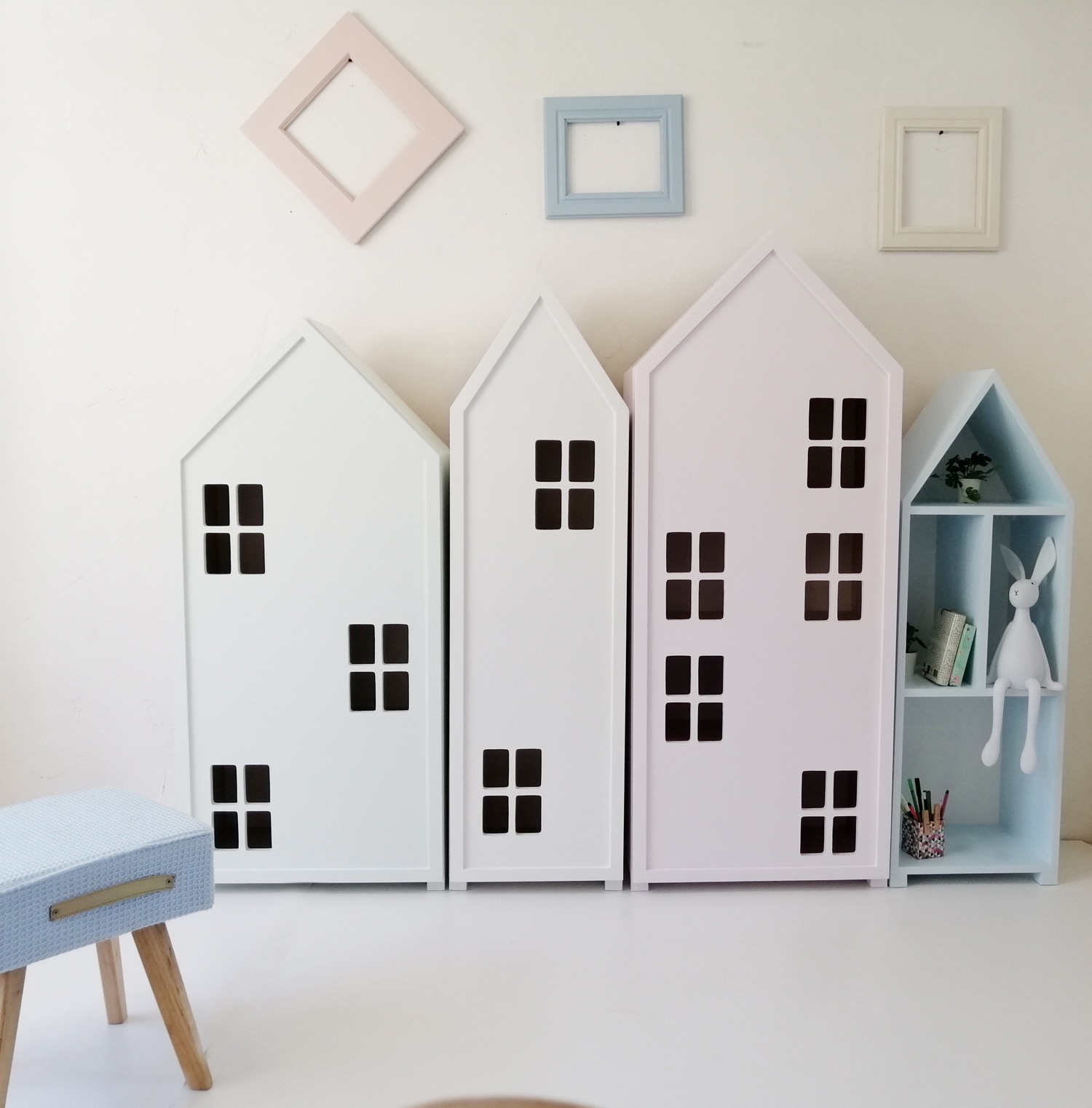Regały dziecięce w kształcie domków, białe i różowe, z kwadratowymi otworami imitującymi okna, ustawione obok siebie, z niebieskim regałem z półkami po prawej stronie, na tle białej ściany z ramkami.