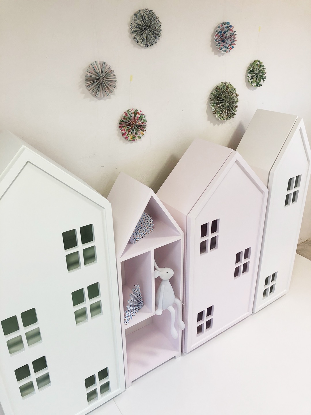 Biało-różowe regały dziecięce w kształcie domków z okienkami, uzupełnione papierowymi dekoracjami na ścianie i pluszowym królikiem w jednej z półek.