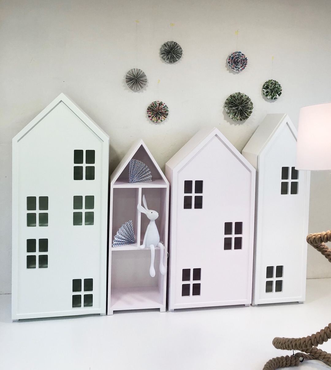 Białe i różowe szafki w kształcie domków z czarnymi oknami, jedna z półkami i pluszowym królikiem w środku, dekoracje papierowe na ścianie, lampa z grubym sznurem w prawym dolnym rogu.