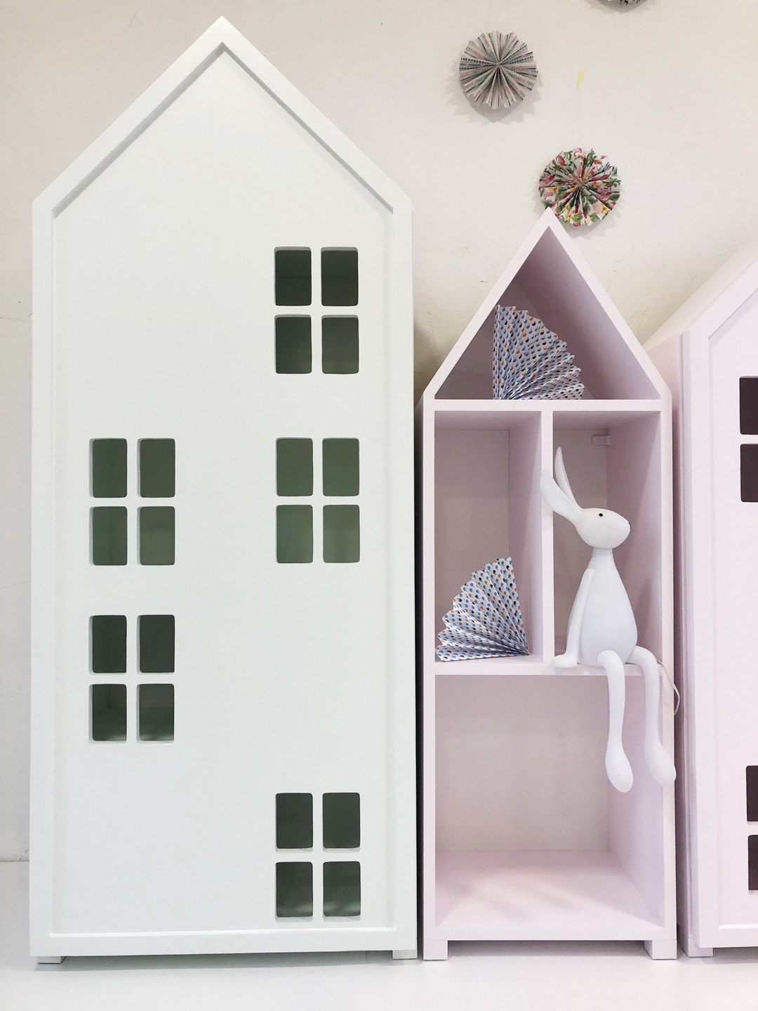 Białe i różowe regały w kształcie domków dla dzieci, z dekoracjami papierowymi i lampką w kształcie królika.