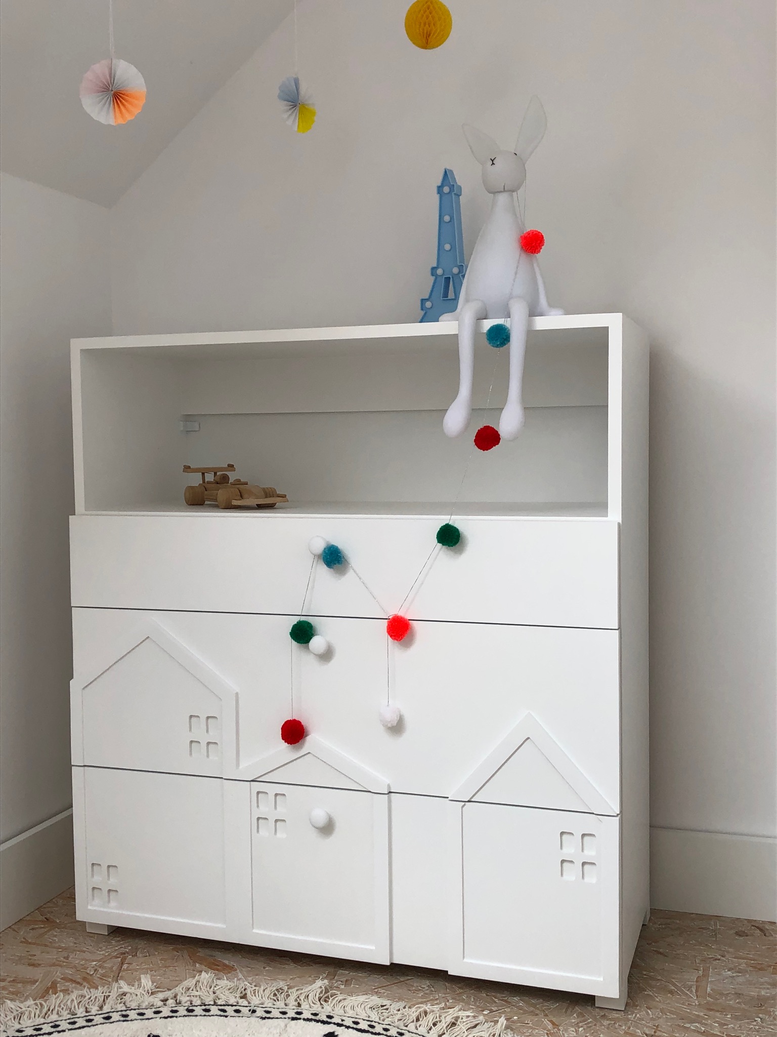 Biała komoda dziecięca z motywem domków, ozdobiona kolorowymi pomponami, na górze leży drewniany samochodzik i siedzi pluszowy królik z wieżą Eiffla w tle.