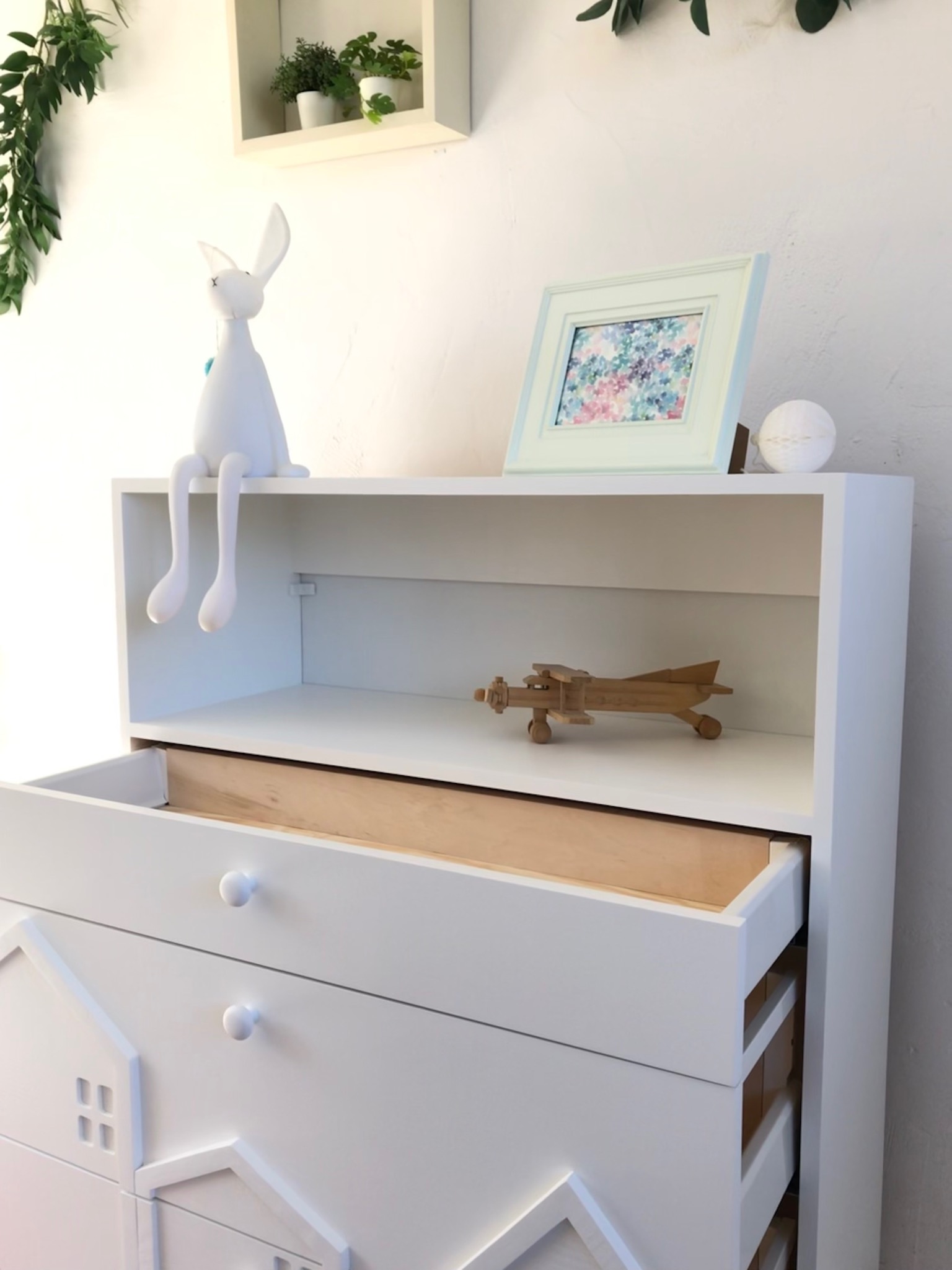 Biała komoda z otwartą szufladą, fronty szuflad zdobione przestrzennymi domkami, na górze półka z drewnianym samolotem, figurką królika i ramką ze zdjęciem.