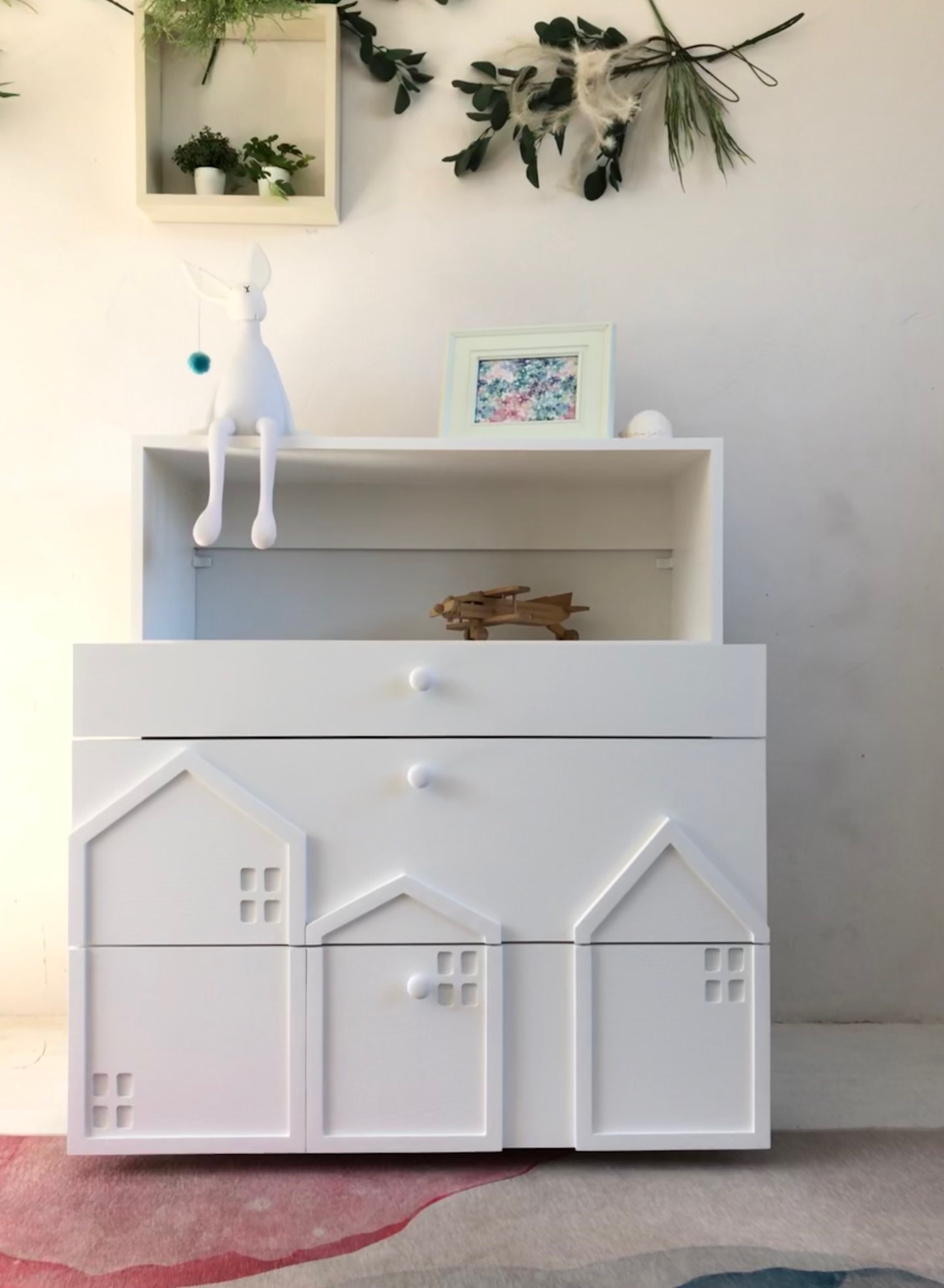 Biała komoda dla dziecka z szufladami w kształcie domków, ozdobiona drewnianym modelem samolotu i pluszowym królikiem, ustawiona na wzorzystym dywanie.