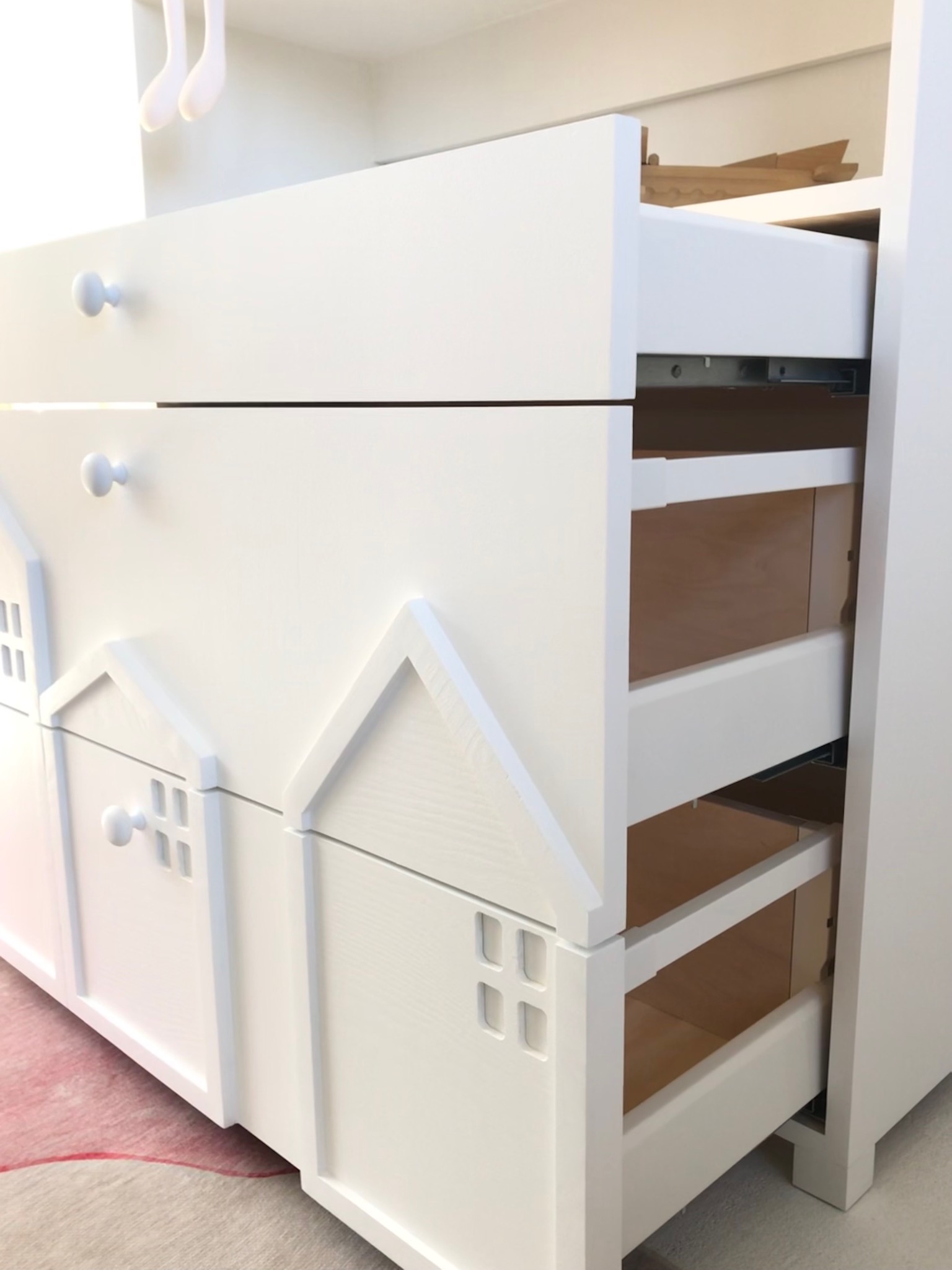 Biała komoda dziecięca z otwartą szufladą, fronty zdobione motywem domków z okienkami i okrągłymi uchwytami.