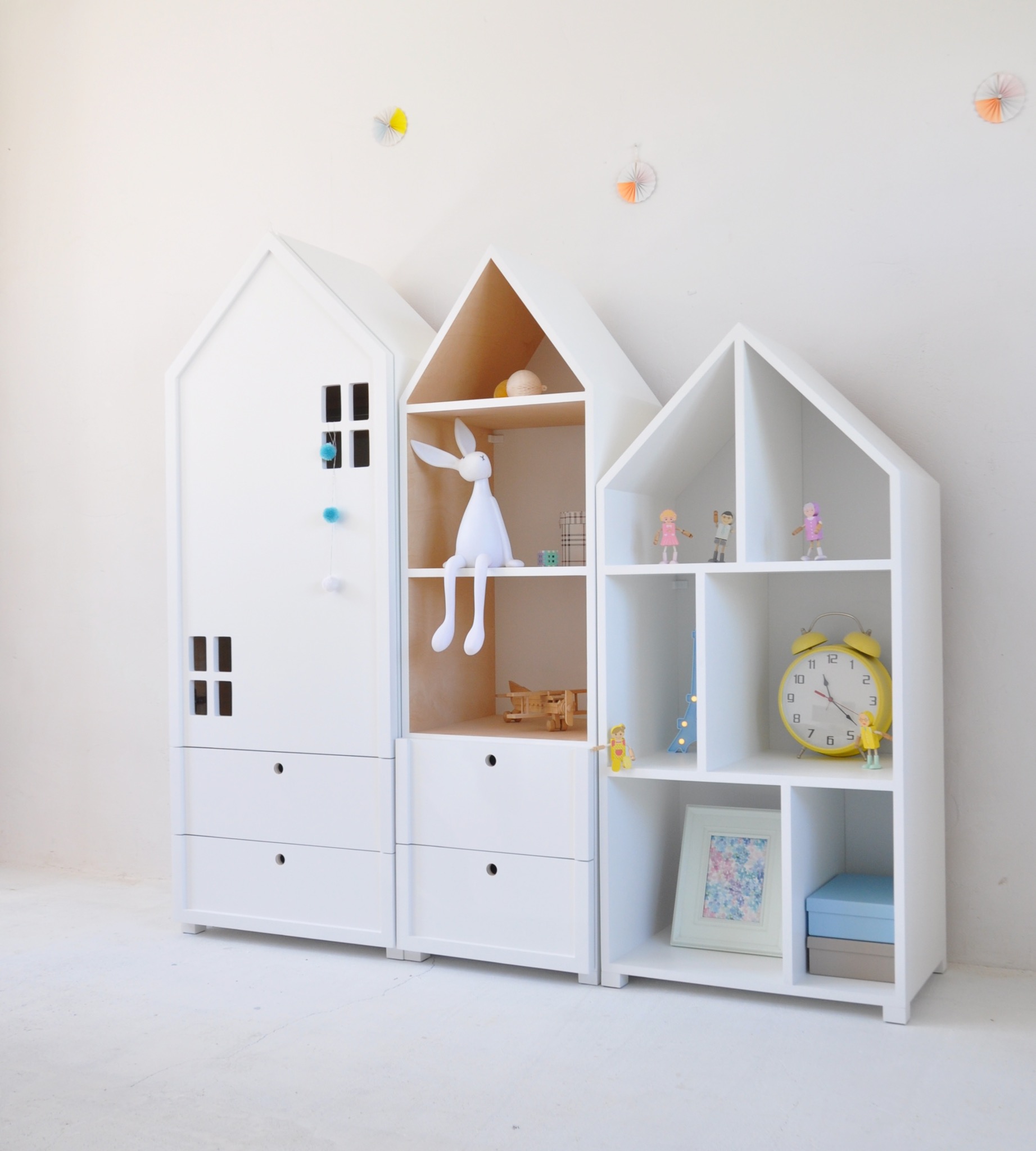 Białe meble dziecięce w kształcie domków z szufladami i półkami, z dekoracjami w postaci zabawek i ozdób, na tle białej ściany.