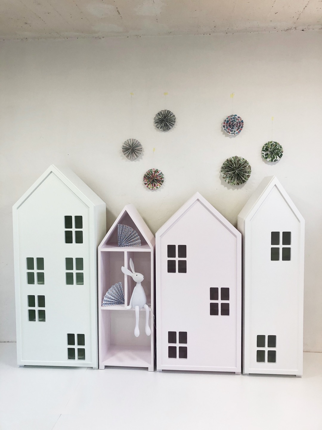 Białe i różowe regały w kształcie domków z oknami, z dekoracjami na ścianie i pluszowym królikiem w środkowym regale.