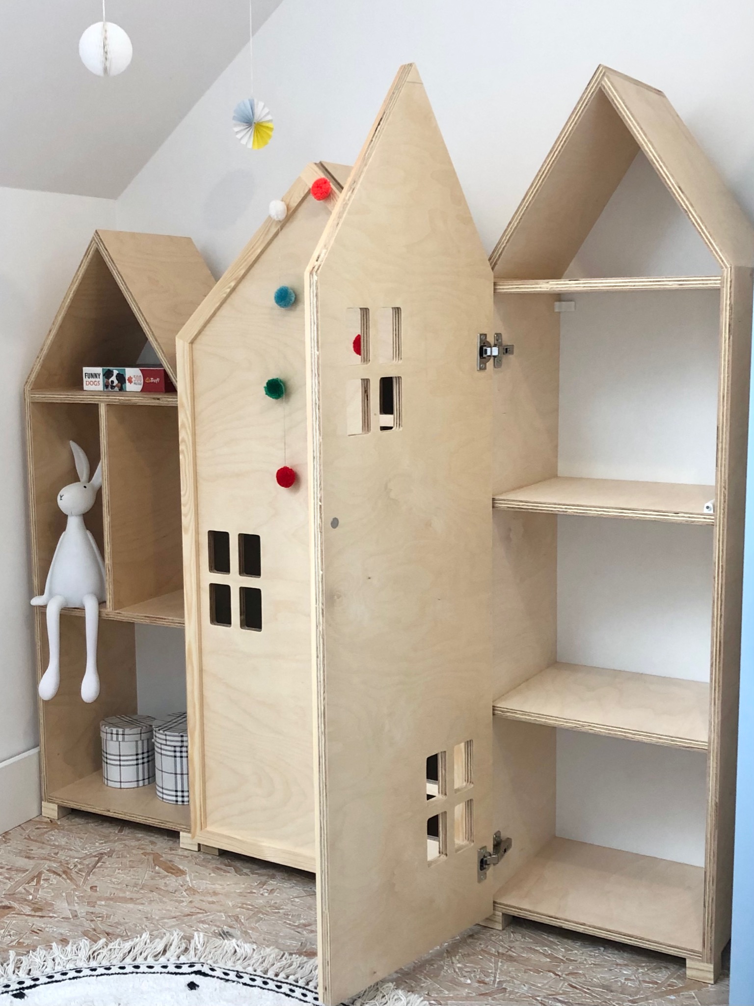 Trzy drewniane regały w kształcie domków dla dzieci, środkowy z otwieranymi drzwiczkami z wyciętymi oknami, ozdobiony kolorowymi pomponami, w tle białe ściany i dekoracje sufitowe.