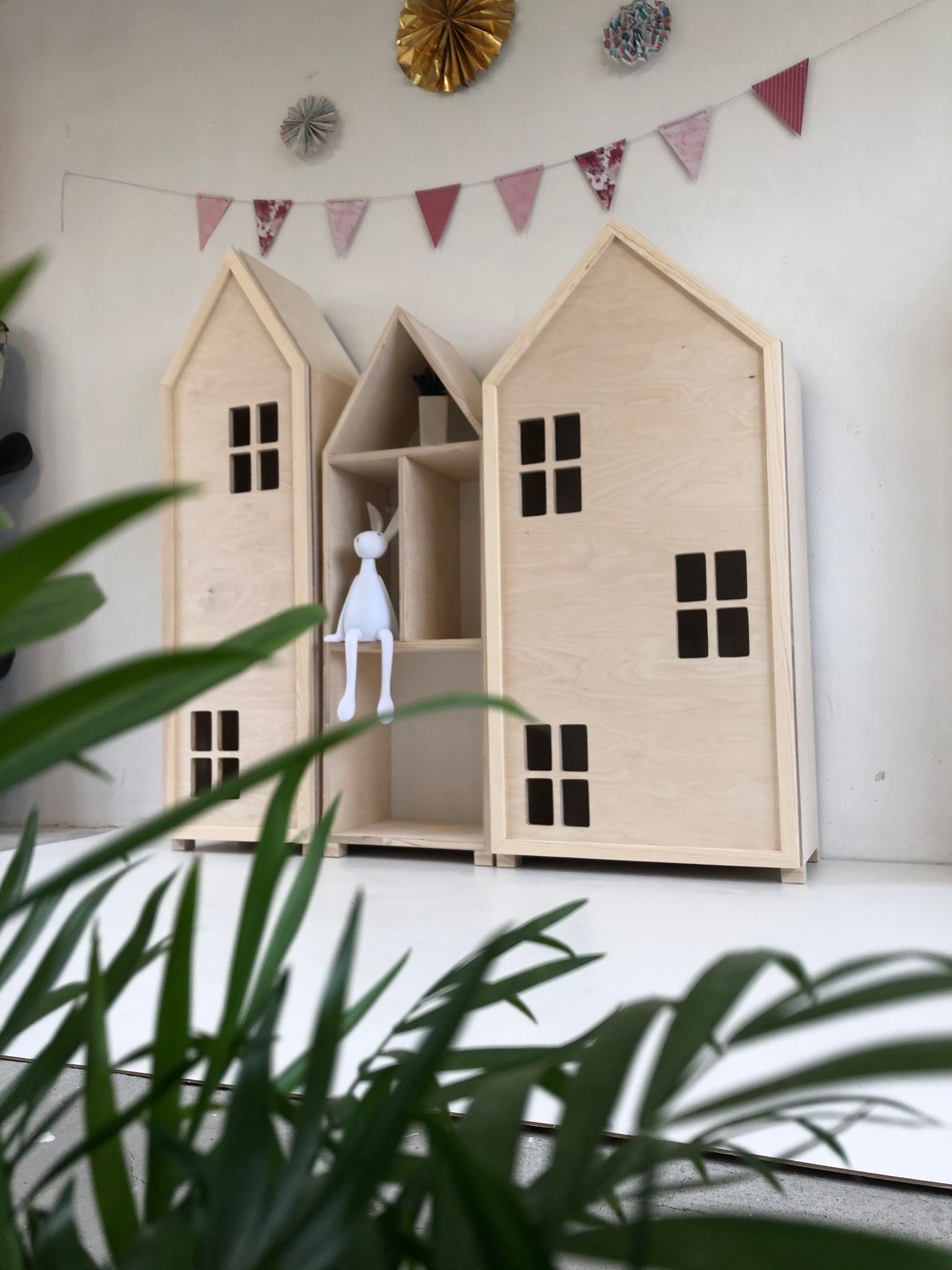 Trzy drewniane regały w kształcie domków, z dekoracyjnymi oknami i półkami, z białym króliczkiem siedzącym na jednej z półek, tło stanowi biała ściana z girlandą z trójkątów i dekoracjami z papieru...