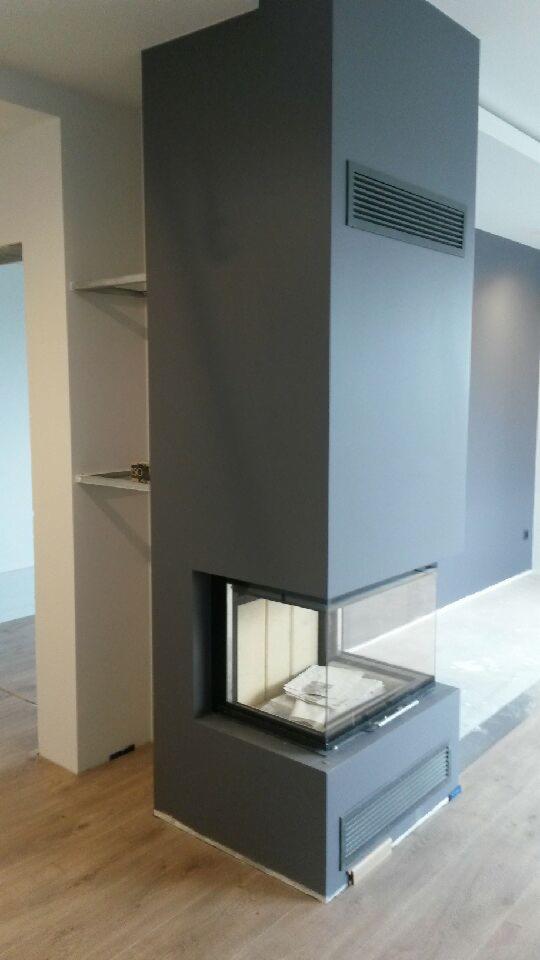 Nowoczesny, szary piec wolnostojący z przeszklonym paleniskiem, wbudowany w ścianę, z widocznymi kratkami wentylacyjnymi i drewnianą podłogą.