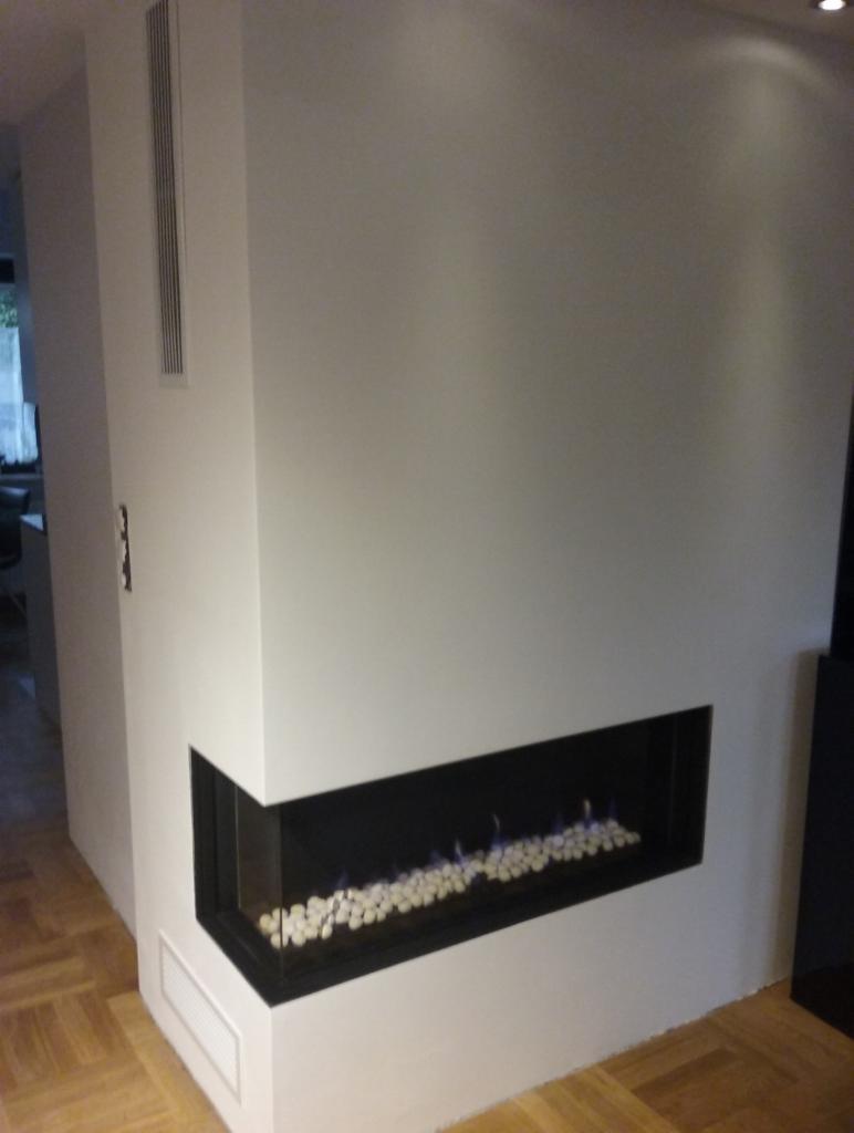 Nowoczesny, narożny biokominek w zabudowie z białych płyt, z paleniskiem wypełnionym białymi kamieniami i delikatnym, niebieskawym płomieniem.