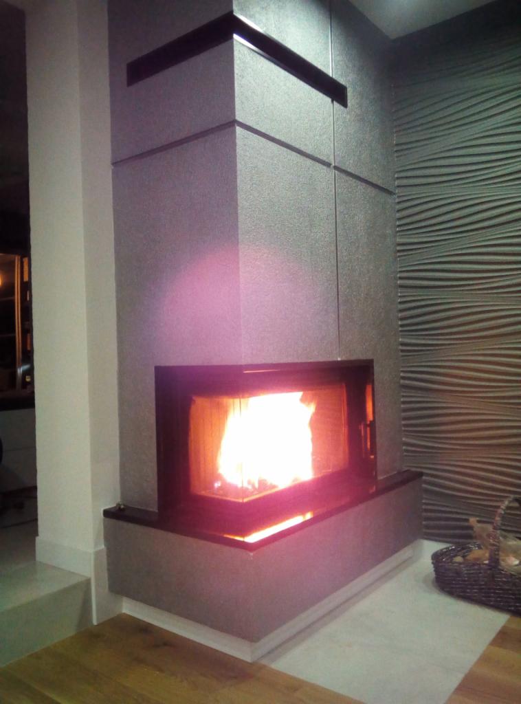 Nowoczesny wkład kominkowy z czarną ramą, zabudowany w szarą ścianę z dekoracyjną fakturą fal, płonący jasnym ogniem.