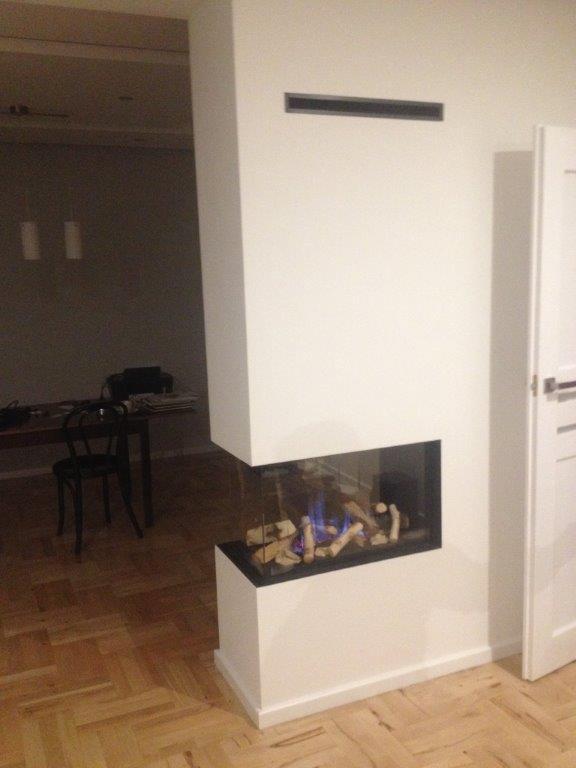 Nowoczesny kominek narożny z szybą, wbudowany w białą ścianę, z widocznym płomieniem i drewnem opałowym, na tle parkietu i częściowo widocznego pomieszczenia z meblami.