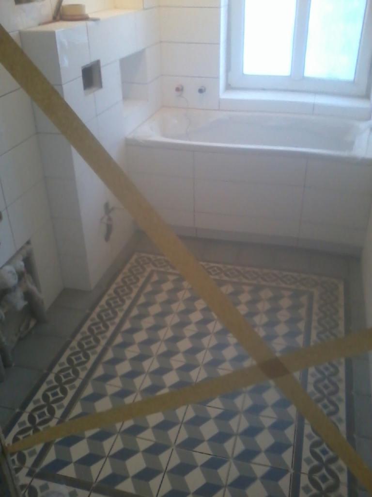 Łazienka w trakcie prac wykończeniowych: białe płytki ścienne, wanna obudowana płytkami, podłoga z wzorzystej, geometrycznej mozaiki w odcieniach bieli, szarości i błękitu; widoczne zabezpieczenia...