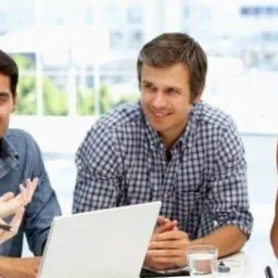 Grupa pięciu osób, różnej narodowości, siedzi przy stole z laptopem, prawdopodobnie podczas lekcji lub spotkania biznesowego, z uśmiechami na twarzach i gestykulacją.