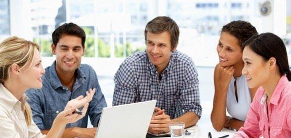 Grupa pięciu osób, różnej narodowości, siedzi przy stole z laptopem, prawdopodobnie podczas lekcji lub spotkania biznesowego, z uśmiechami na twarzach i gestykulacją.