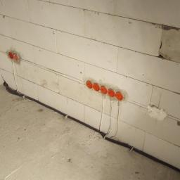 AMW Projekt - Ściana z betonu komórkowego z zamontowanymi puszkami elektrycznymi z pomarańczowymi pokrywkami, prowadzone kable w peszlu, widoczne zwoje kabli w tle.